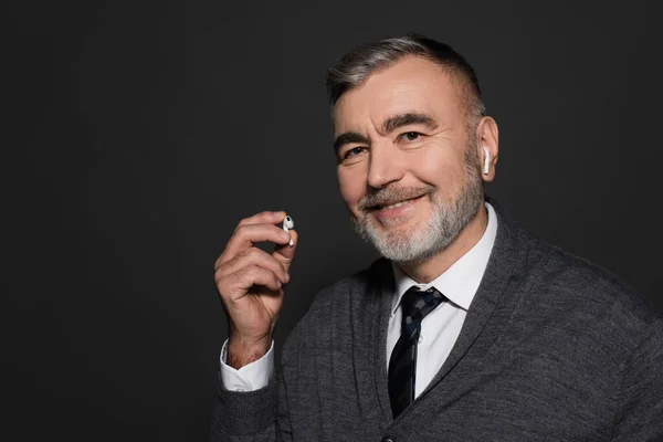 Senior man holding earphone while listening music and smiling at camera isolated on dark grey — Stock Photo