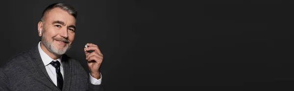 Cheerful senior man holding earphone while listening music isolated on black, banner — Stock Photo