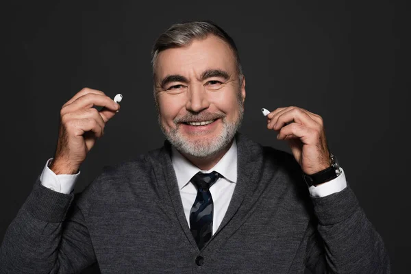 Happy senior man in jumper and tie holding earphones isolated on dark grey — Stockfoto