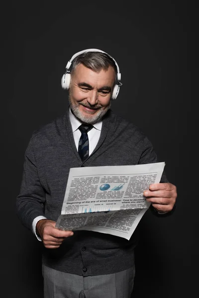 Happy senor man in jumper, tie and headphones reading newspaper on dark grey — Stock Photo