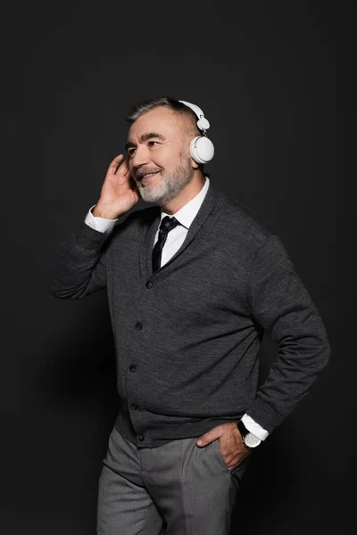 Старший стильний чоловік стоїть з рукою в кишені, слухаючи музику в навушниках на темно-сірому — стокове фото