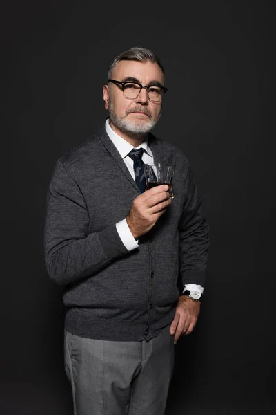 Stylish and confident senior man standing with hand on hip and glass of whiskey on dark grey — стоковое фото