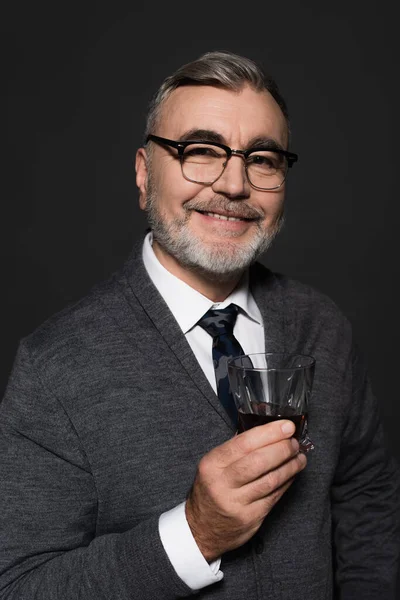 Positive senior man in eyeglasses holding glass of whiskey while looking at camera isolated on dark grey — стоковое фото