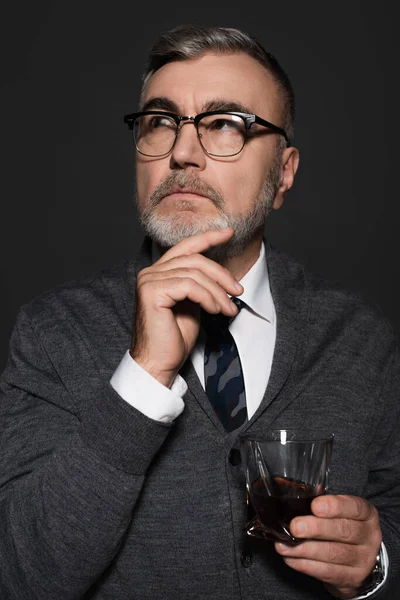 Thoughtful senior man with glass of whiskey holding hand near face while looking away isolated on dark grey — стоковое фото