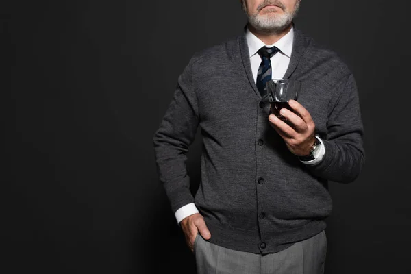 Cropped view of stylish man with glass of whiskey standing with hand in pocket on dark grey — стоковое фото