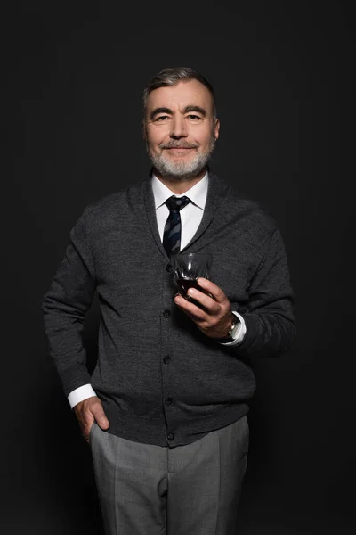 Grey-haired man standing with glass of whiskey and hand in pocket while smiling at camera on dark grey — Stockfoto