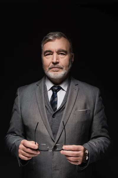 Senior bearded businessman in grey suit holding eyeglasses and looking at camera isolated on black — стоковое фото