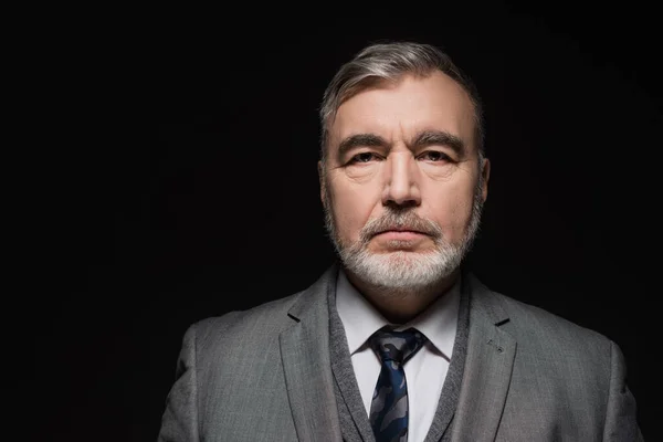 Senior bearded man in grey blazer and tie looking at camera isolated on black — Stockfoto