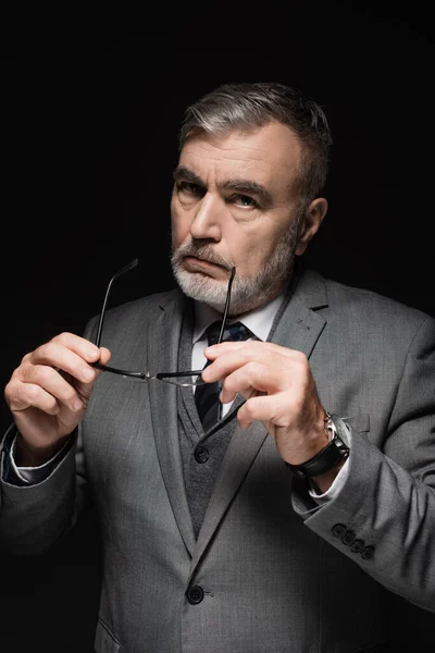 Confident senior businessman in blazer holding eyeglasses and looking at camera isolated on black — стоковое фото