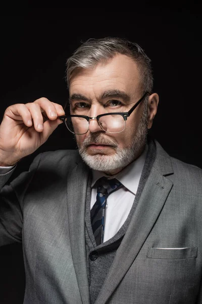 Serious senior businessman adjusting eyeglasses while looking at camera isolated on black — Fotografia de Stock