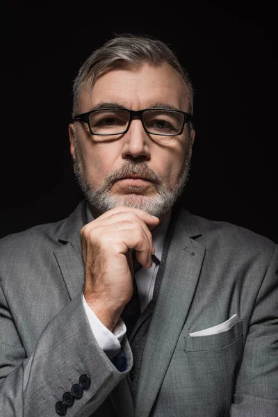 Senior pensive businessman in eyeglasses touching chin while looking at camera isolated on black — Stockfoto