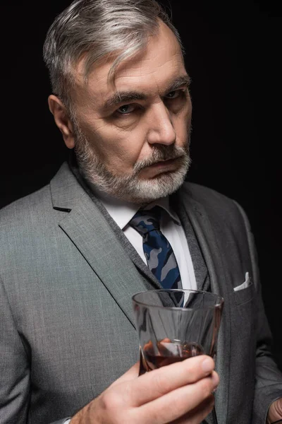 Strict businessman in blazer and tie looking at camera near glass of whiskey isolated on black — Stock Photo