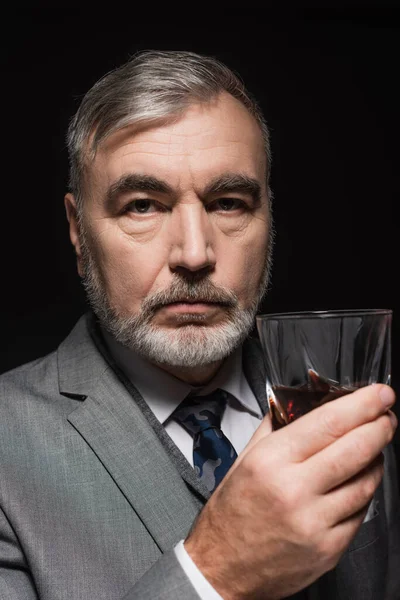 Senior bearded man in blazer and tie holding glass of whiskey isolated on black — стоковое фото