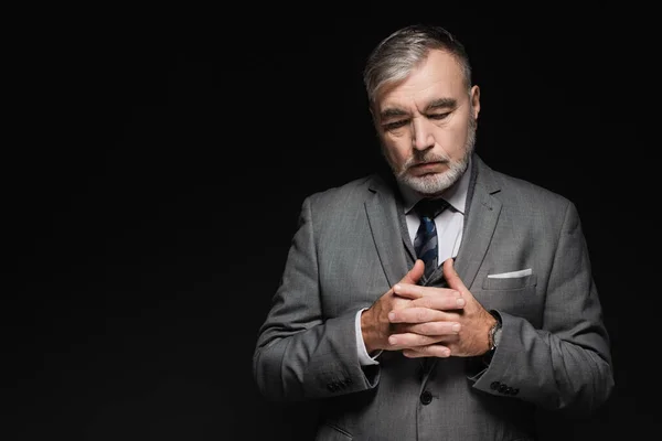 Thoughtful senior man in blazer standing with clasped hands isolated on black — Photo de stock