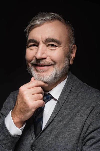 Positive senior man smiling at camera while touching chin isolated on black — стоковое фото