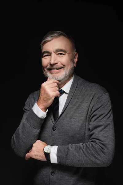 Happy senior man in jumper and tie smiling at camera isolated on black — стоковое фото