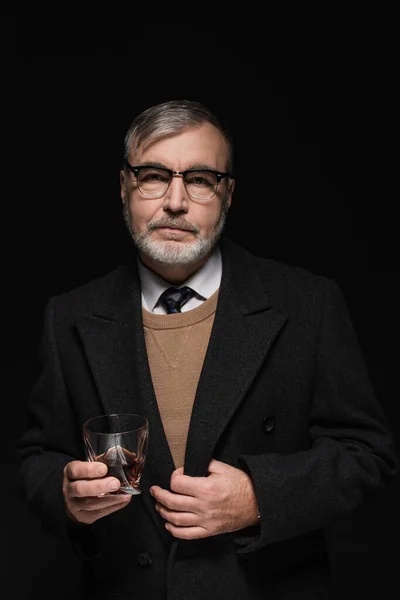 Senior bearded man in coat and eyeglasses holding glass of whiskey isolated on black — Photo de stock