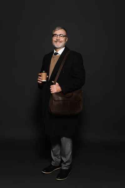 Full length view of pleased senior man in black coat standing with leather bag on dark grey — стоковое фото