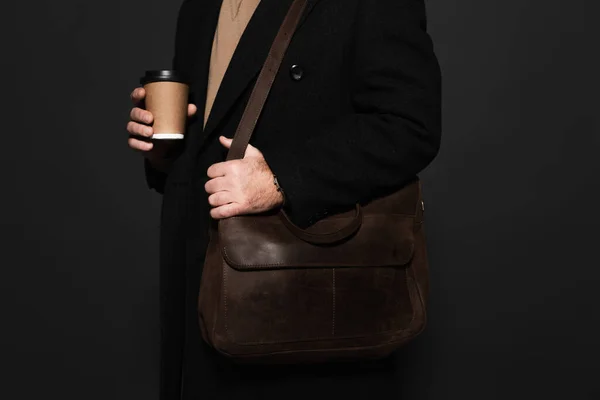 Partial view of trendy man with coffee to go and leather bag isolated on black — стоковое фото