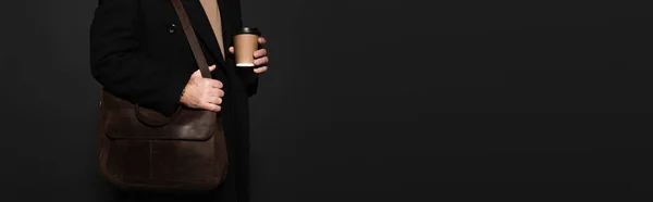 Partial view of man in coat standing with paper cup and leather bag isolated on black, banner — Stock Photo