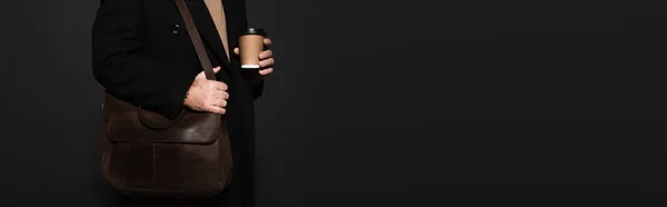 Partial view of man in coat standing with paper cup and leather bag isolated on black, banner — стоковое фото