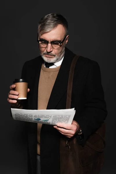 Trendy senior man with leather bag and paper cup reading newspaper isolated on dark grey — Stockfoto