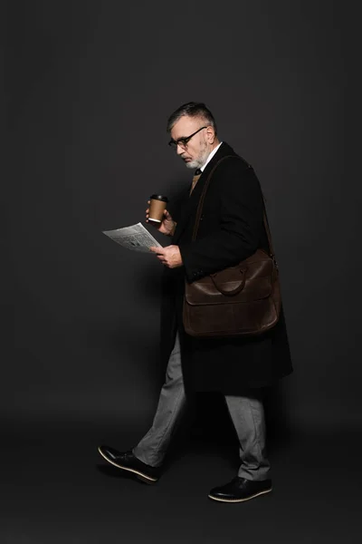 Full length view of trendy senior man with coffee to go and newspaper walking on dark grey — Stockfoto