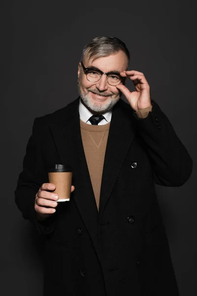 Happy man in stylish coat holding paper cup and adjusting eyeglasses isolated on dark grey — Fotografia de Stock