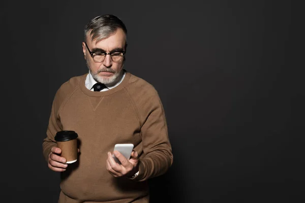 Elderly man with coffee to go looking at mobile phone on dark grey — Stockfoto