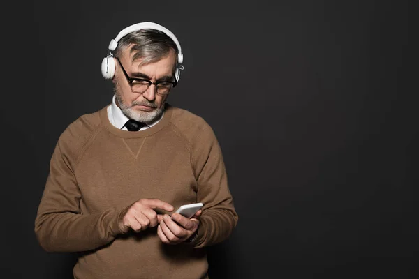 Senior man in beige jumper and headphones pointing at mobile phone on dark grey — Stockfoto