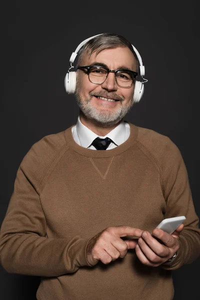 Cheerful senior man in headphones looking at camera while holding smartphone isolated on black — стоковое фото