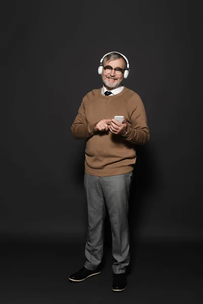 Повнометражний вид на стильного старшого чоловіка в навушниках, що тримає смартфон і посміхається на камеру на темно-сірому — стокове фото