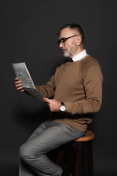 Stylish senior man in eyeglasses reading newspaper while sitting on stool on dark grey — Foto stock