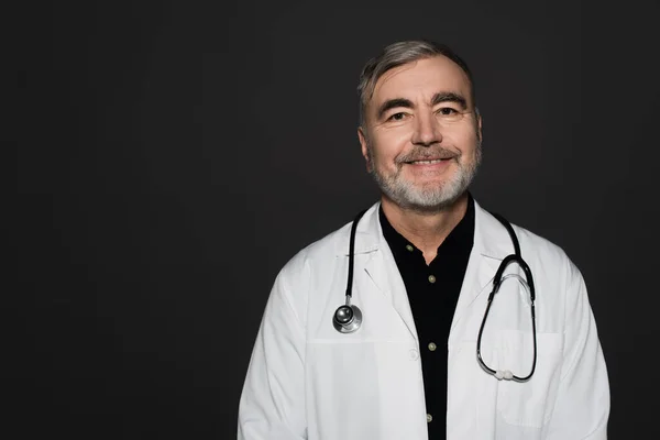 Smiling bearded doctor with stethoscope looking at camera isolated on black — Foto stock