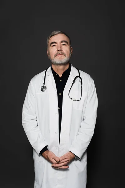 Bearded doctor in white coat standing with clasped hands and looking at camera isolated on dark grey - foto de stock