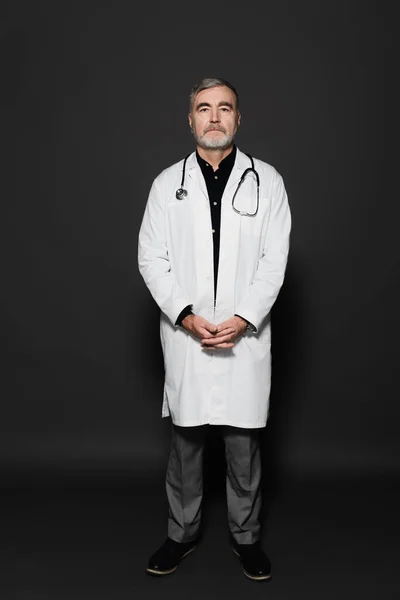 Full length view of on senior physician in white coat looking at camera on dark grey — Foto stock