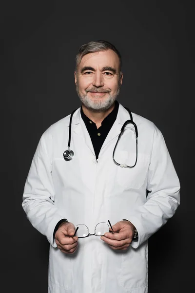 Positive bearded physician holding eyeglasses and smiling at camera isolated on black — Stock Photo