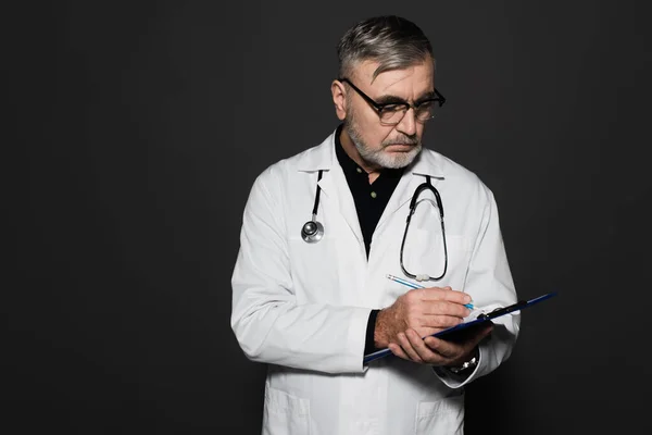 Старший лікар в окулярах і білому пальто, що пише на кишені ізольовано на чорному — стокове фото