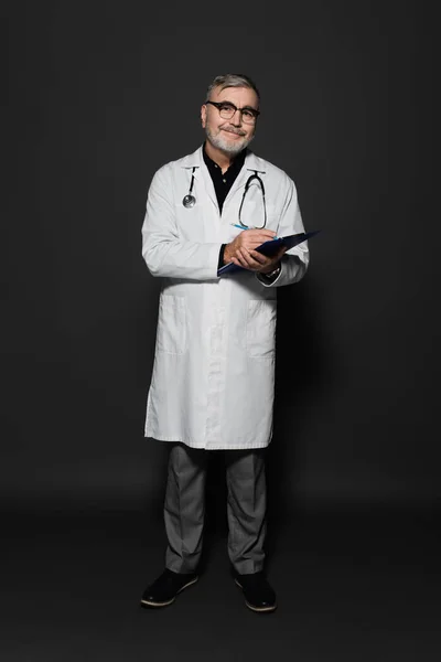 Full length view of happy senior doctor in white coat writing on clipboard on dark grey — Stock Photo