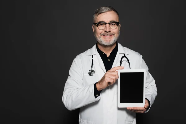 Joyful senior doctor in white coat and eyeglasses holding digital tablet with blank screen isolated on black — Foto stock