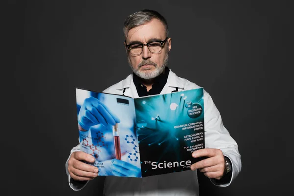 Senior doctor in eyeglasses reading science magazine isolated on dark grey — Stock Photo