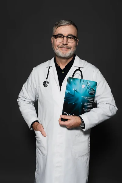 Smiling physician in eyeglasses standing with magazine and hand in pocket of white coat isolated on dark grey — Stock Photo