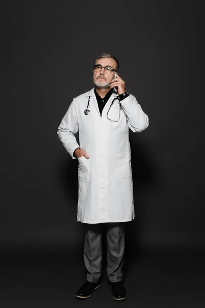 Full length view of senior doctor standing with hand in pocket and talking on smartphone on dark grey — Stock Photo