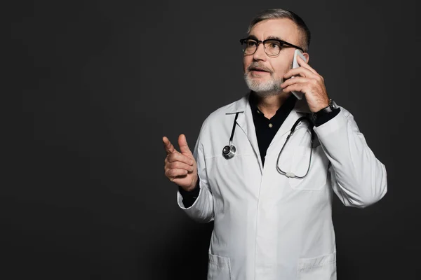 Bearded doctor looking away and pointing with finger while talking on cellphone on dark grey - foto de stock