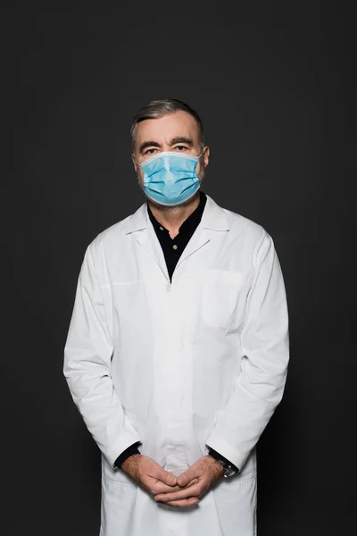 Senior physician in white coat and medical mask looking at camera isolated on dark grey — Stock Photo