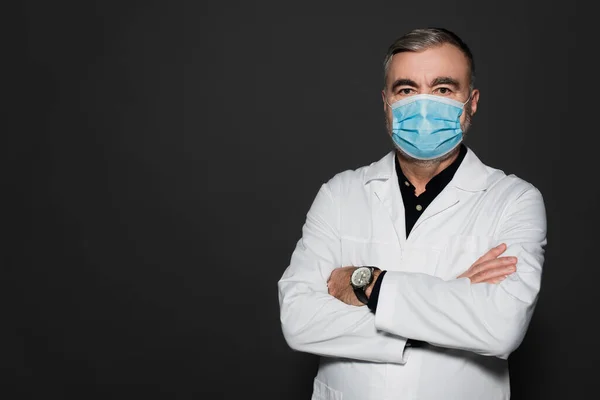 Doctor in white coat and medical mask standing with crossed arms and looking at camera isolated on dark grey — Stockfoto