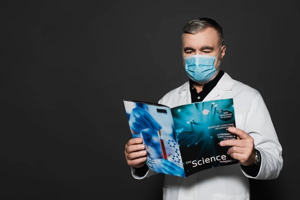 Senior physician in medical mask reading science magazine isolated on dark grey — Stock Photo