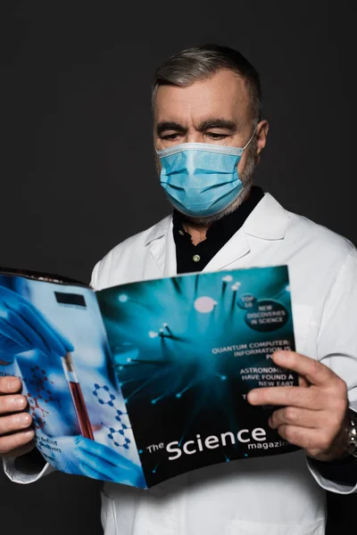 Physician in medical mask reading science magazine isolated on dark grey — Stock Photo