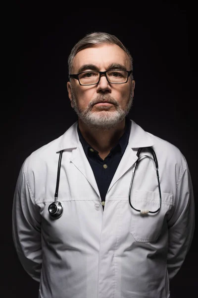 Senior bearded doctor with stethoscope looking at camera isolated on black — Stock Photo