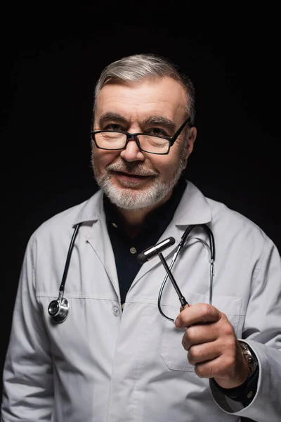Positive neurologist in white coat and eyeglasses holding reflex malleus isolated on black — стоковое фото